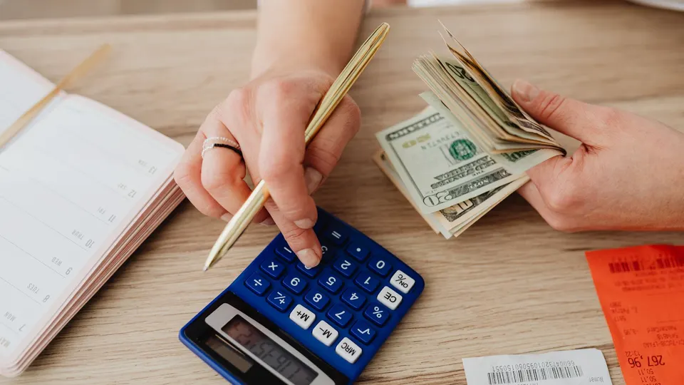 A hand using a blue calculator beside U.S. dollar bills